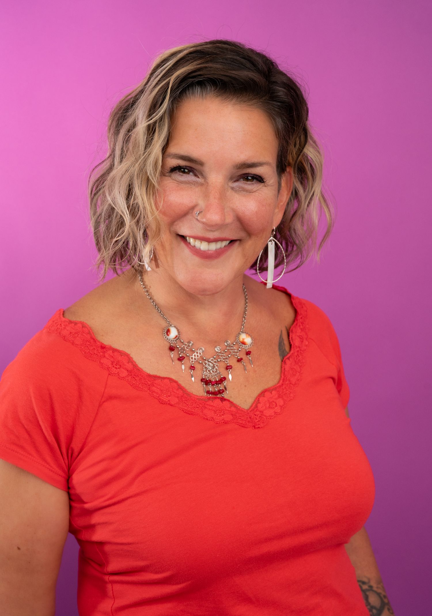 Smiling woman with wavy blonde hair wearing a red top and statement necklace poses against a vibrant purple background.