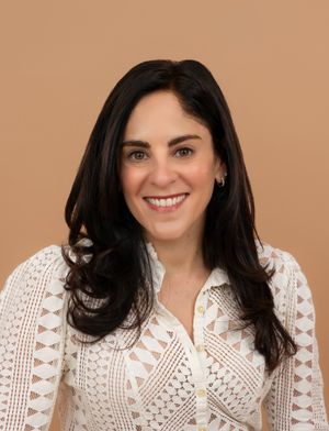 Smiling woman with dark hair wearing a white lace blouse against a warm beige background.
