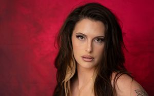Young woman with long dark hair poses against a vibrant red background with a tattoo visible on her shoulder.