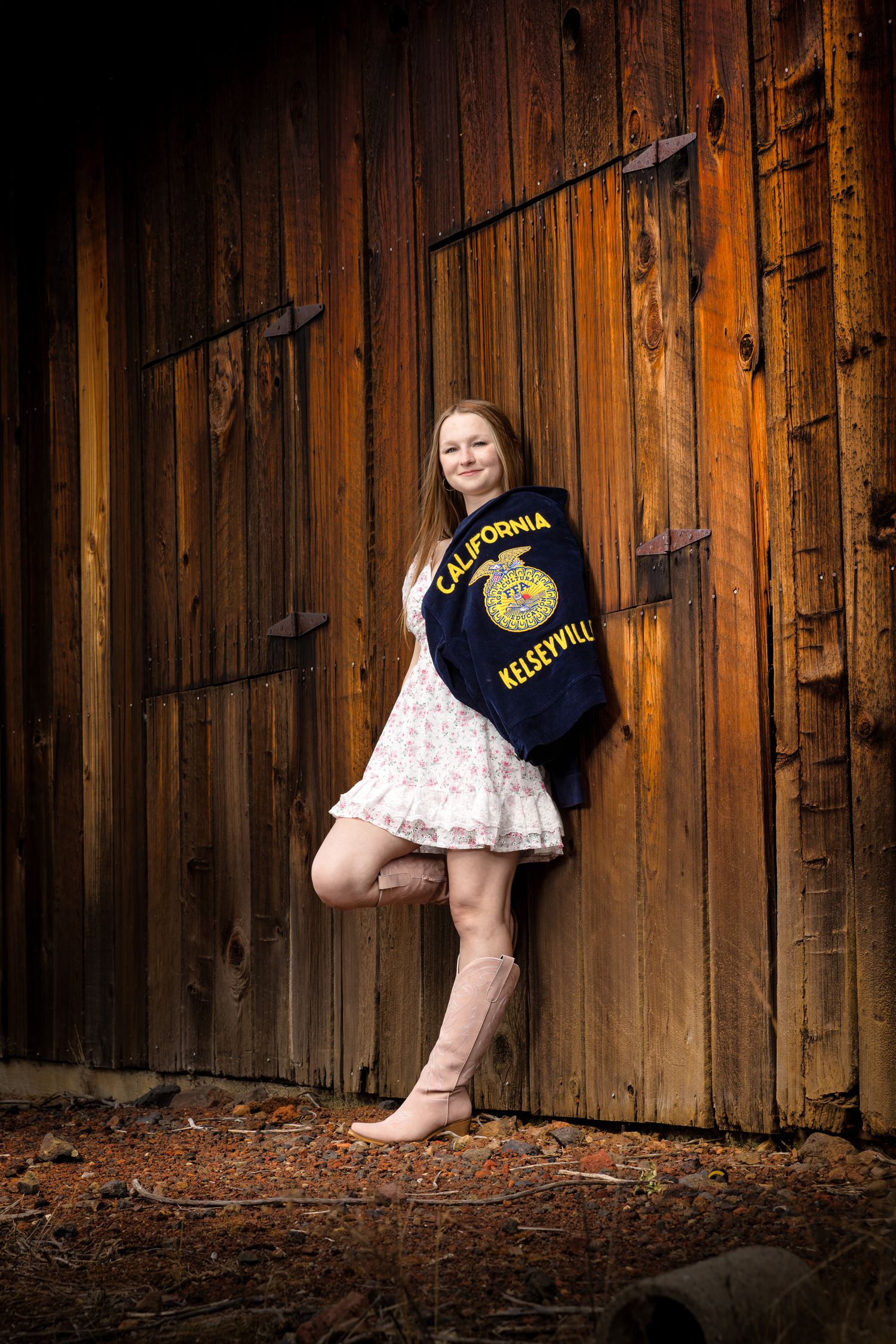Young woman in floral dress and pink boots leans against rustic barn wall wearing a California Kelseyville FFA jacket.
