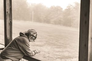 Woman with glasses leaning on a wooden railing, gazing at a misty green field in a sepia-toned outdoor photo.