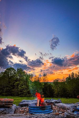 Vibrant sunset sky over a stone fire pit with roaring flames surrounded by lush green trees.