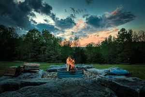 Twilight campfire glowing in a stone ring pit under dramatic cloudy sky with forest backdrop.