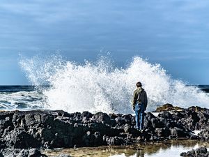 Person standing on rocky shore watching massive ocean waves crash dramatically against the coastline under a cloudy blue sky.