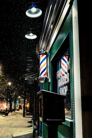 Barber shop exterior at night with classic striped barber poles glowing in the snow on a city street.