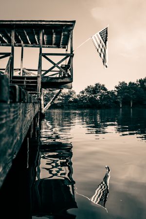 Sepia-toned American flag flying from a rustic wooden dock over calm reflective lake waters with trees in background.
