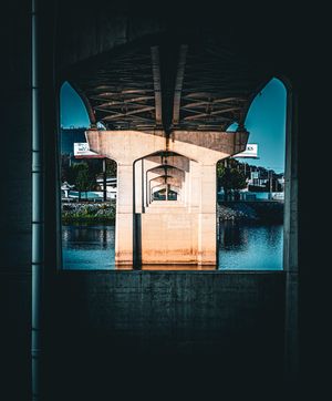 Symmetrical view under a concrete bridge over calm water, with arched supports and blue sky visible beyond.