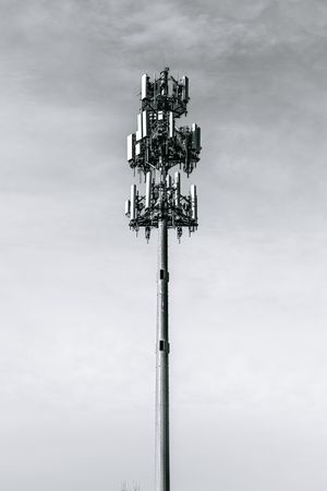 Black and white photo of a tall cellular communication tower with multiple antennas against a cloudy sky.