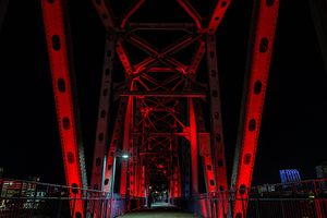 Red-lit steel truss bridge glowing dramatically against a dark night sky with city lights in the background.