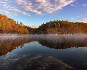 Misty autumn lake reflecting colorful fall foliage and blue sky with clouds at sunrise.
