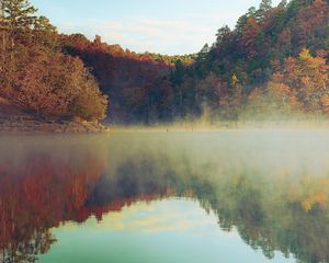 Serene misty lake surrounded by vibrant autumn trees reflecting warm fall colors in calm water.