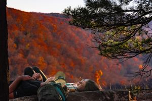 Two hikers resting on a rock ledge overlooking a vibrant autumn forest with fiery red and orange foliage.