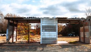 Rustic open-sided metal shed with corrugated walls surrounded by autumn trees under a cloudy sky in a rural setting.