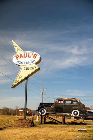 Vintage car displayed on a ramp beside Paul's Body Shop retro star-shaped sign under a blue sky.
