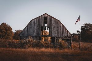 Weathered wooden barn in autumn field with American flag against gray sky.