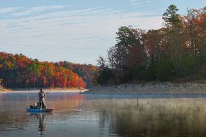 Angler fishing from a small boat on a calm lake surrounded by vibrant autumn foliage in morning mist.