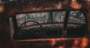 Rusty abandoned vintage car interior with weathered steering wheel and corroded dashboard viewed through broken window.