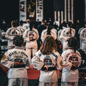 Young martial artists in white gis with championship patches stand at attention before an American flag at a karate tournament.