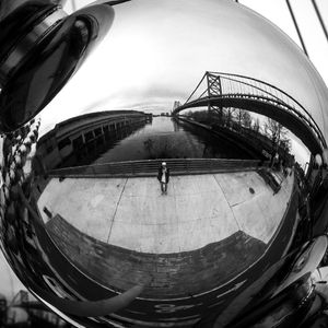 Black and white fisheye reflection of a person standing on a waterfront promenade with a suspension bridge in the background.