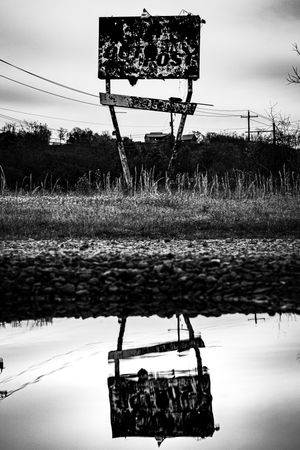 Black and white photo of a weathered roadside sign reflected in a puddle, set against an overcast sky.