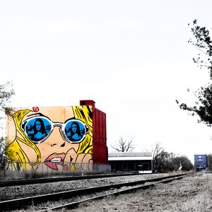 Street art mural of a pop art blonde woman wearing blue sunglasses near railroad tracks.