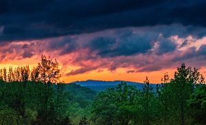 Dramatic sunset sky with vivid orange and red clouds over silhouetted trees and rolling mountain hills.