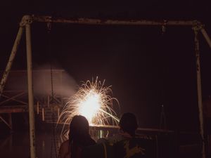 Two people watching bright fireworks sparkle against a dark night sky near a dock.