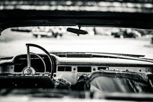 Black and white interior view of a vintage classic car dashboard with steering wheel and rearview mirror.