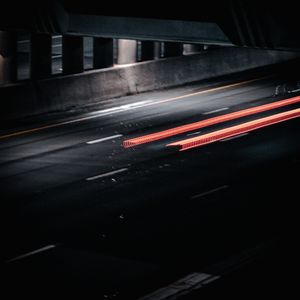 Long exposure night photography captures red light trails streaking under a dark urban overpass on a wet city road.