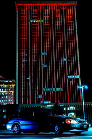 A vintage car parked at night in front of a red-lit skyscraper in an urban cityscape.