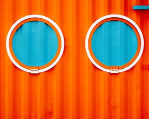 Two round porthole windows with white frames on a vibrant orange corrugated metal wall revealing turquoise blue interior.