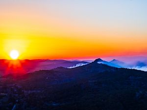Stunning mountain sunset with vibrant orange and yellow sky, misty blue peaks, and sun rays over a scenic landscape.