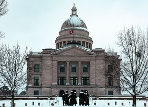 Snow-covered state capitol building with dome and flag, bare winter trees, and silhouetted figures in foreground.