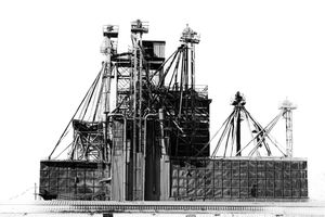 Black and white industrial facility with large cranes and steel framework structures against a white sky.