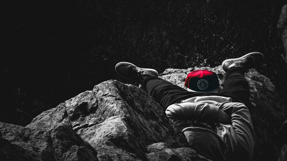 Person in white jacket and red hat resting on dark rocky terrain in a dramatic black and white outdoor scene.