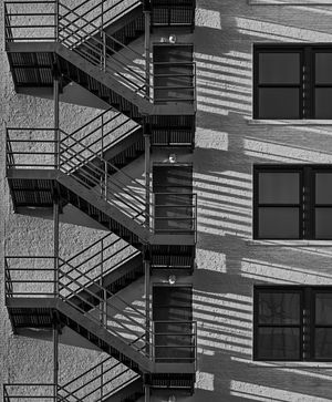 Black and white photo of zigzagging metal fire escape stairs casting dramatic shadows on a building exterior wall.