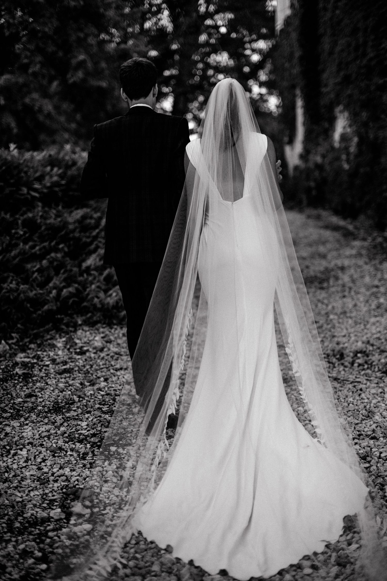 Black and white photograph of Beth and Matt walking together, her long veil trailing behind.