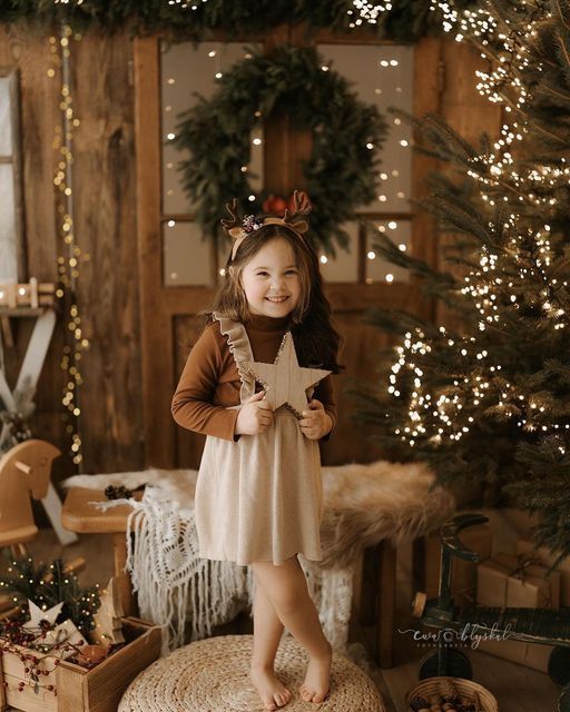 Young girl in reindeer headband holding a wooden star amid Christmas trees and twinkling lights in a festive holiday setting.