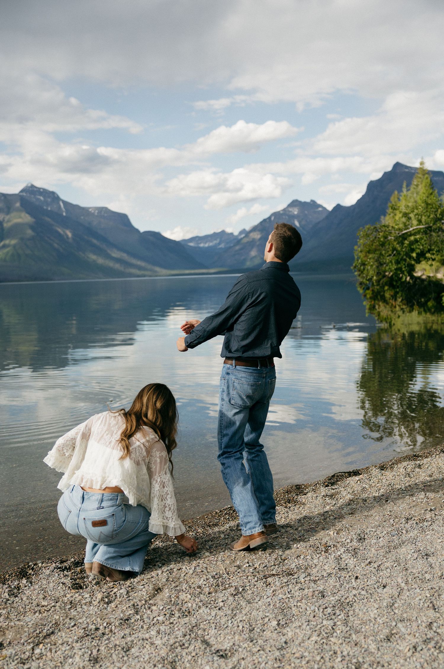 Couple skipping stones at mountain lake shore with stunning reflective water and peaks behind.
