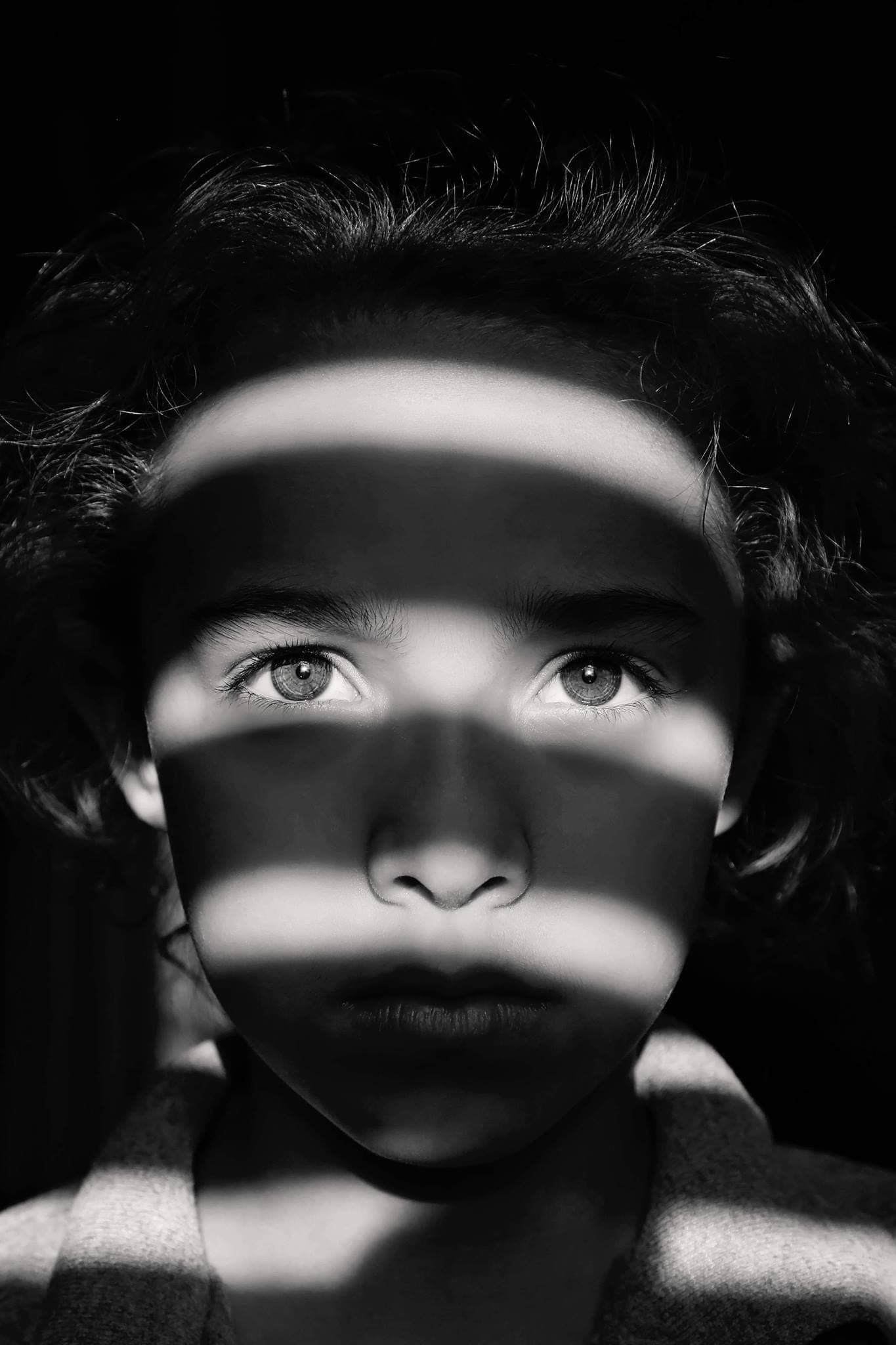Black and white portrait of a child with striking eyes and dark curly hair, with stripes of light and shadow across the face.