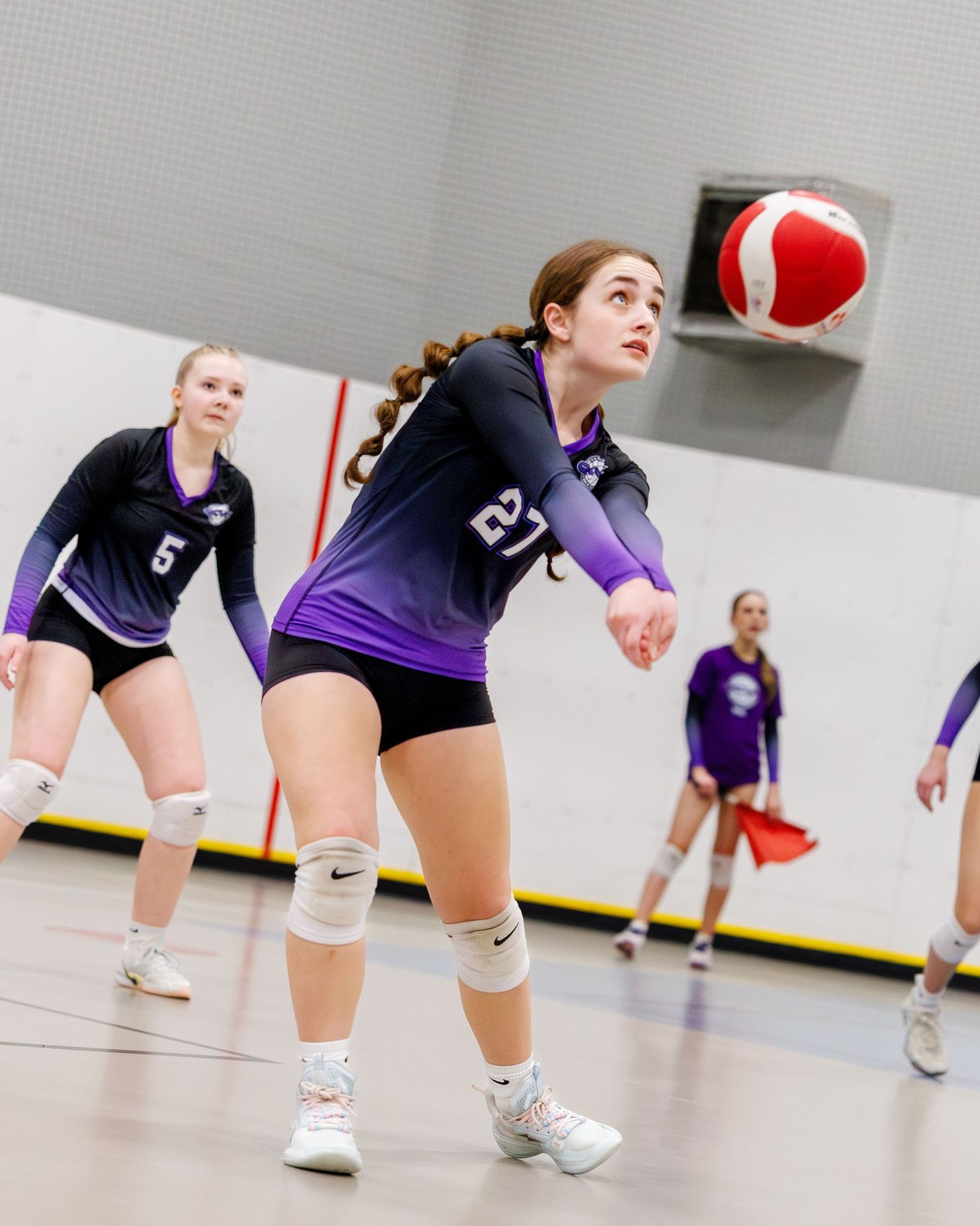 Volleyball player #27 in purple uniform bumps ball during indoor match.