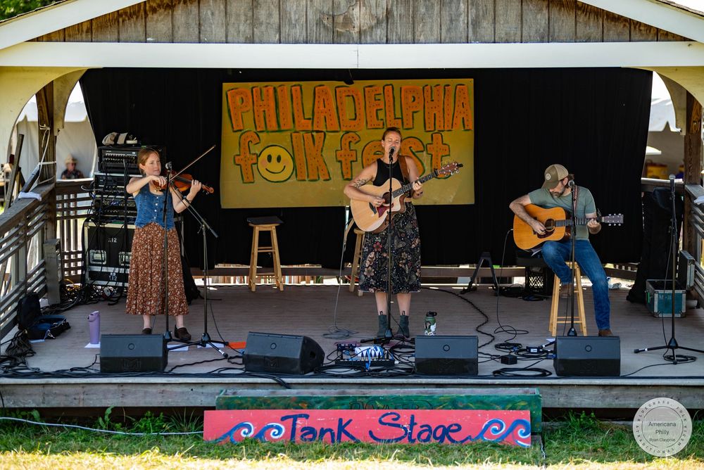 Three musicians perform on the Tank Stage at Philadelphia Folk Fest, featuring violin, vocals, and acoustic guitar outdoors.