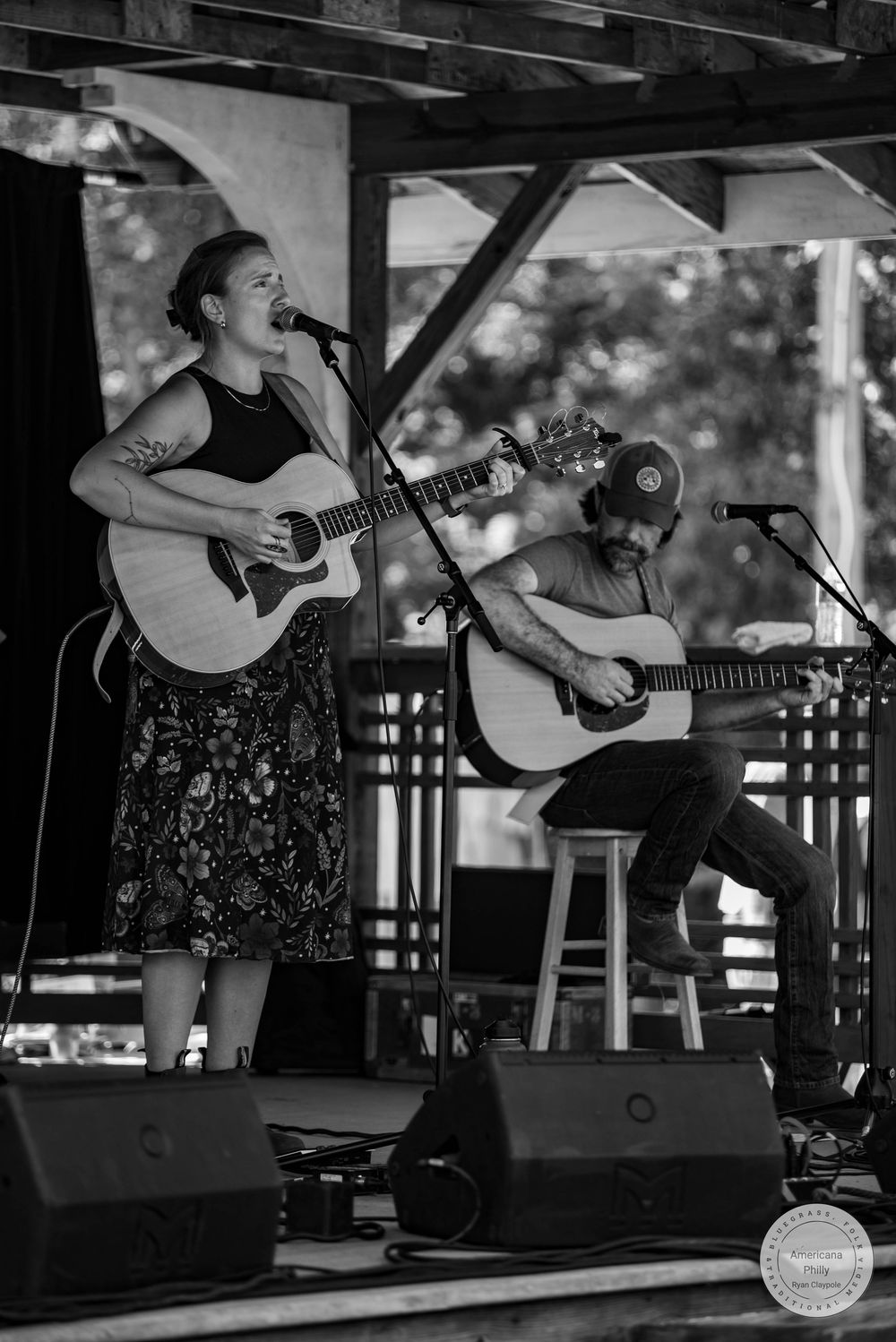 Two musicians perform acoustic guitars on an outdoor stage, woman singing into mic in black floral skirt, black and white photo.