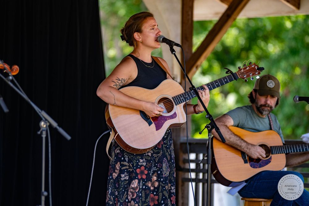 Woman singing and playing acoustic guitar on outdoor stage with male guitarist performing beside her.