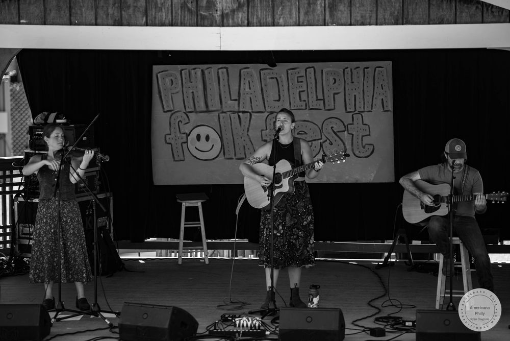 Three musicians perform on stage at Philadelphia Folk Fest, with two women and a man playing acoustic guitars and violin.