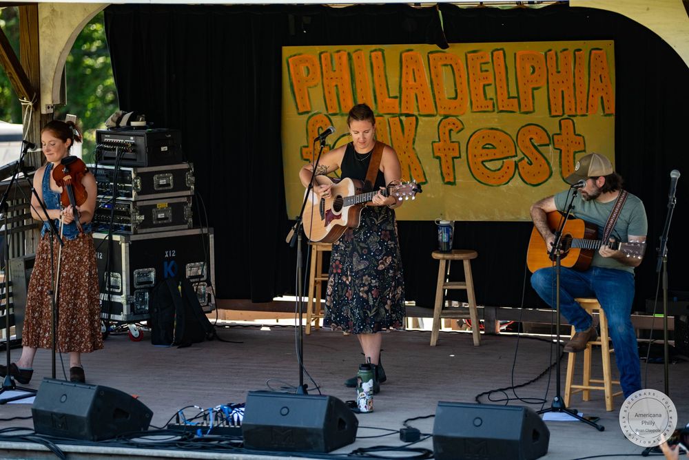 Three musicians perform live at Philadelphia Folk Fest outdoor stage, two guitarists and a violinist in colorful summer setting.