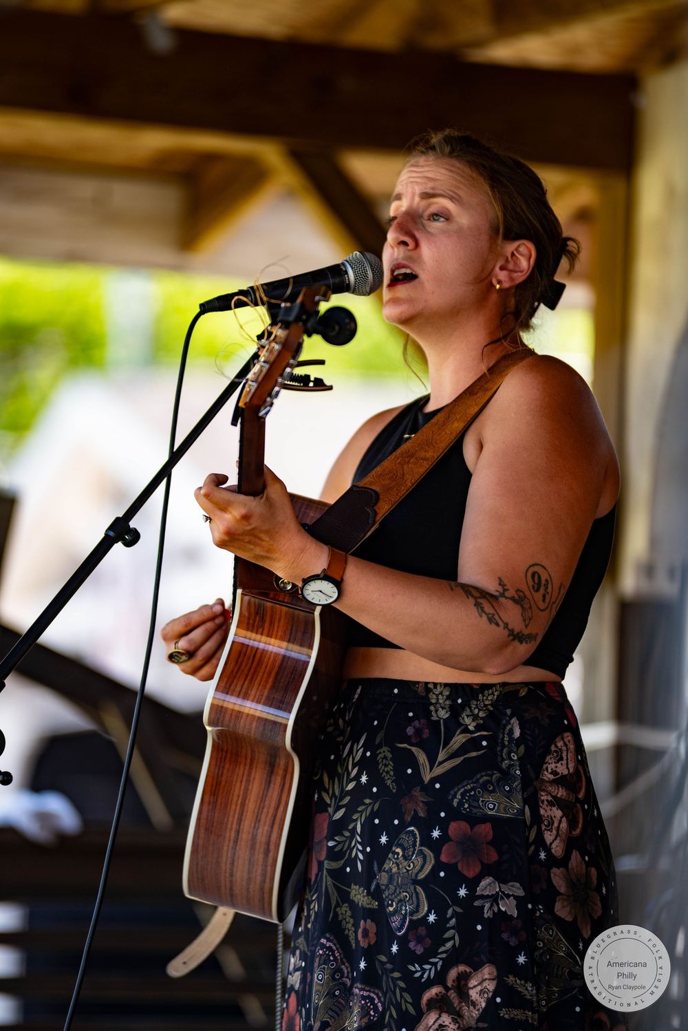 Woman with tattoos plays acoustic guitar and sings into microphone at outdoor music festival.