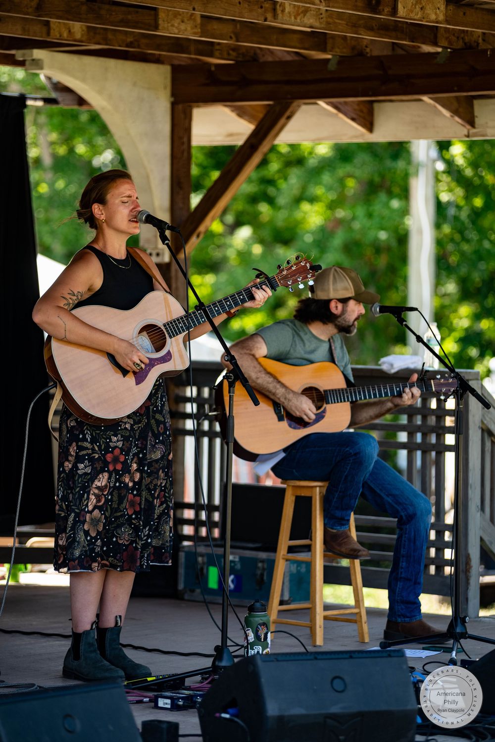 Two musicians perform acoustic guitars outdoors at a pavilion stage, woman singing into mic wearing floral skirt.