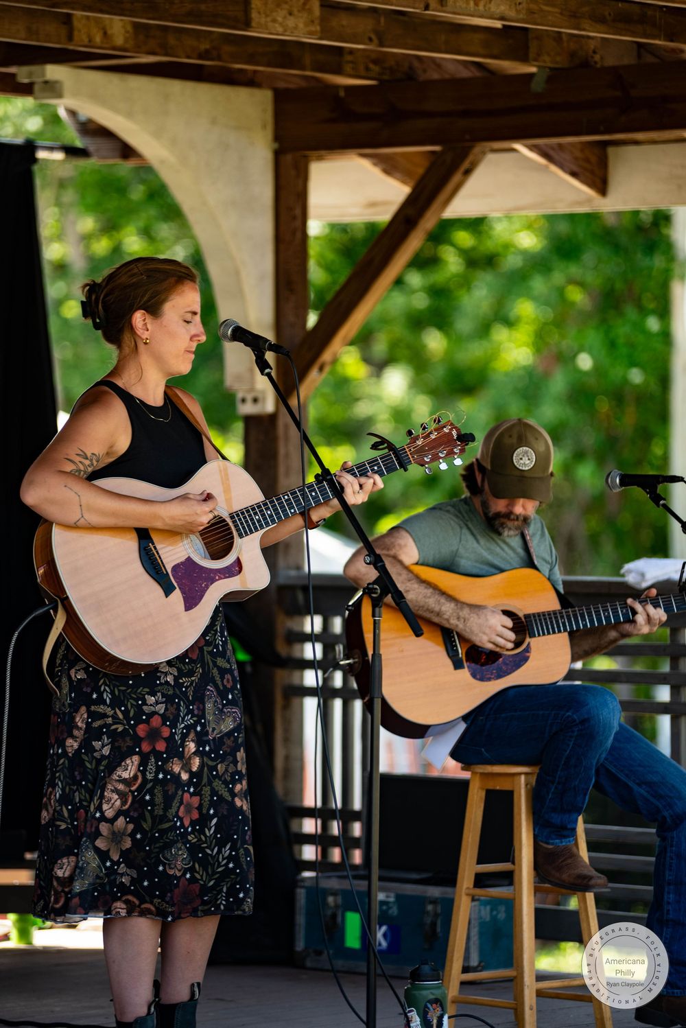 Two musicians playing acoustic guitars on an outdoor stage, a woman singing at a microphone and a man seated on a stool.