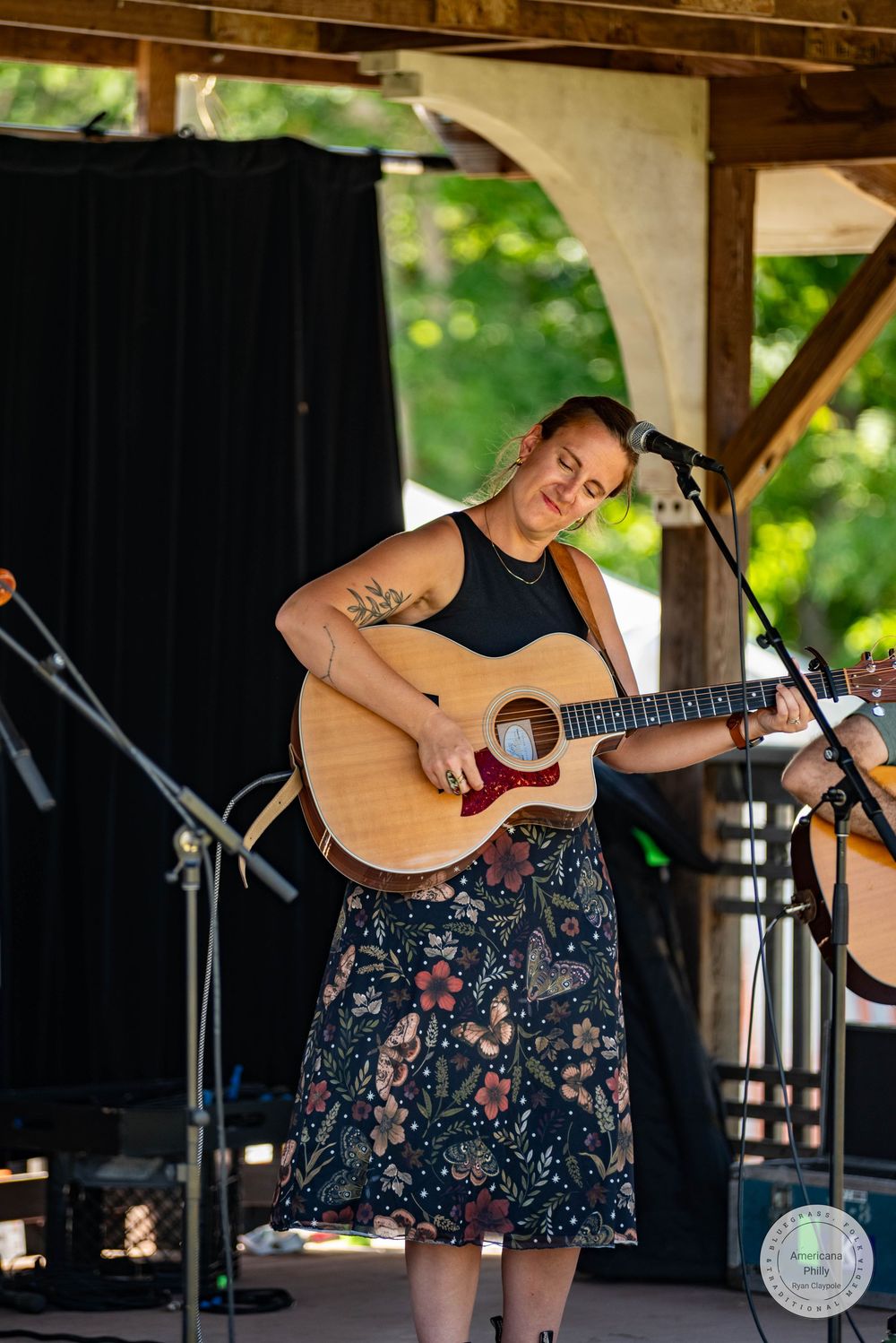 Woman playing acoustic guitar on outdoor stage wearing floral skirt and black top, performing at summer music festival.